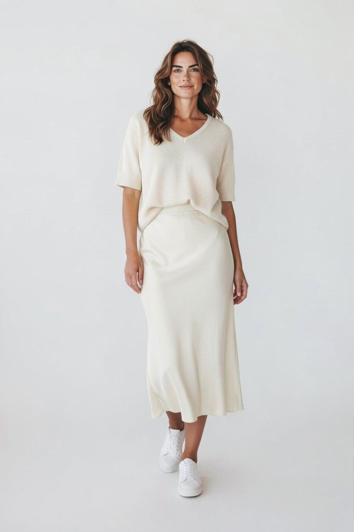 A woman with long wavy hair smiles softly against a light background, wearing the ROSE & CROWN Cashmere V Neck Short Sleeve Sweater and a matching midi skirt, paired with white sneakers.
