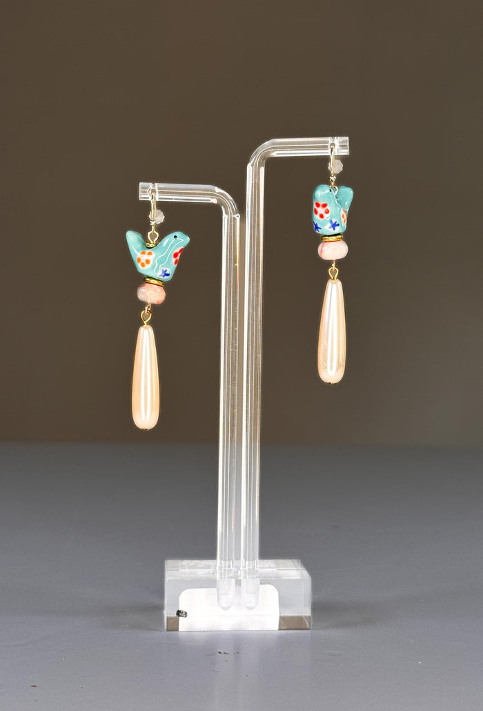 The SANDRINE FRANCE STUDIO - GEMMA EARRINGS feature turquoise ceramic bird charms with painted flowers, beige round beads, and long teardrop-shaped pearl dangles on gold hooks, shown against a white background.