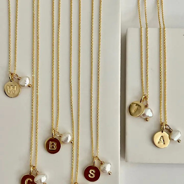 The SANDRINE FRANCE STUDIO TYPWRITER INITIAL NECKLACE "E" features a gold-plated, handmade round pendant engraved with an "E" and paired with a freshwater pearl charm, presented on a delicate chain.