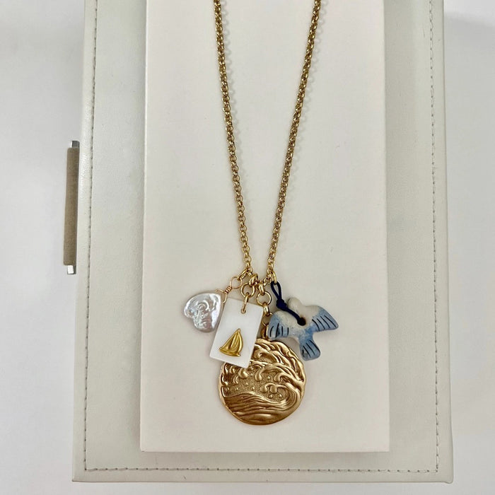 The SANDRINE FRANCE STUDIO WREN NECKLACE is a handmade gold necklace from NYC featuring four charms—a white shell, a rectangular gold sailboat tag, a gold wave pendant, and a blue-white fish—on a white jewelry stand.