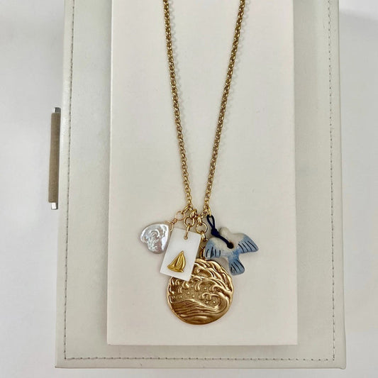 The SANDRINE FRANCE STUDIO WREN NECKLACE is a handmade gold necklace from NYC featuring four charms—a white shell, a rectangular gold sailboat tag, a gold wave pendant, and a blue-white fish—on a white jewelry stand.