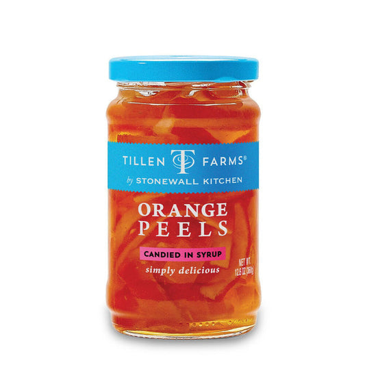A jar of Stonewall Kitchen - Tillen Farms Candied Orange Peels is placed on a marble counter next to a cocktail glass, sliced oranges, and a filled carafe. A jigger, cocktail strainer, and spoon complete this vegan-friendly citrusy treat preparation scene.