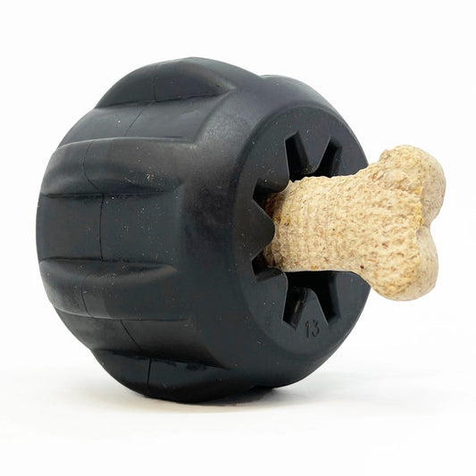 The SODA PUP TREAT HOLDER MAGNUM GEAR (BLACK) is a tough tire-shaped dog chew toy made from natural black rubber, shown holding a tan bone-shaped treat in its center against a white background.