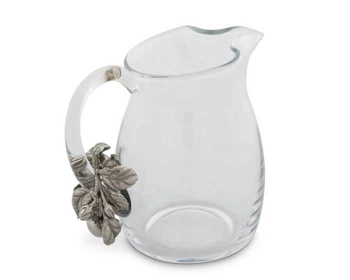 The VAGABOND HOUSE - APPLE GLASS PITCHER from VAGABOND HOUSE features a clear glass body with a curved spout and an ornate handle decorated with silver-toned leaves and acorns, blending traditional craftsmanship into a unique, elegant piece of functional tableware.