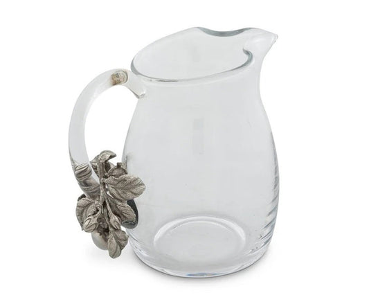 The VAGABOND HOUSE - APPLE GLASS PITCHER from VAGABOND HOUSE features a clear glass body with a curved spout and an ornate handle decorated with silver-toned leaves and acorns, blending traditional craftsmanship into a unique, elegant piece of functional tableware.