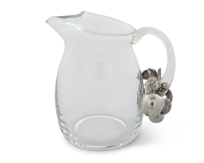 The VAGABOND HOUSE - APPLE GLASS PITCHER from VAGABOND HOUSE features a clear glass body with a curved spout and an ornate handle decorated with silver-toned leaves and acorns, blending traditional craftsmanship into a unique, elegant piece of functional tableware.