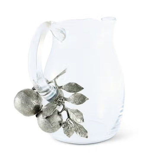 The VAGABOND HOUSE - ORANGE BLOOMS GLASS PITCHER features a clear glass design with a distinctive handle adorned in luxury-grade pewter, showcasing metallic textured oranges and leaves, creating an elegant contrast to the simple transparency of the pitcher.