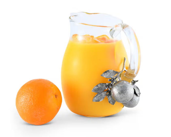 The VAGABOND HOUSE - ORANGE BLOOMS GLASS PITCHER features a clear glass design with a distinctive handle adorned in luxury-grade pewter, showcasing metallic textured oranges and leaves, creating an elegant contrast to the simple transparency of the pitcher.