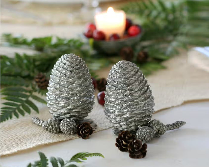 Two detailed silver pine cones stand upright on a white background, resembling the VAGABOND HOUSE - PEWTER PINE CONESALT AND PEPPER SET by VAGABOND HOUSE, with small pine needle segments visible at their bases.