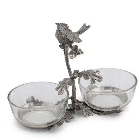 The VAGABOND HOUSE - SONG BIRD DOUBLE CONDIMENT BOWL features two glass bowls on an elegant metal stand adorned with ornate leaves and branches, topped by a pewter songbird.