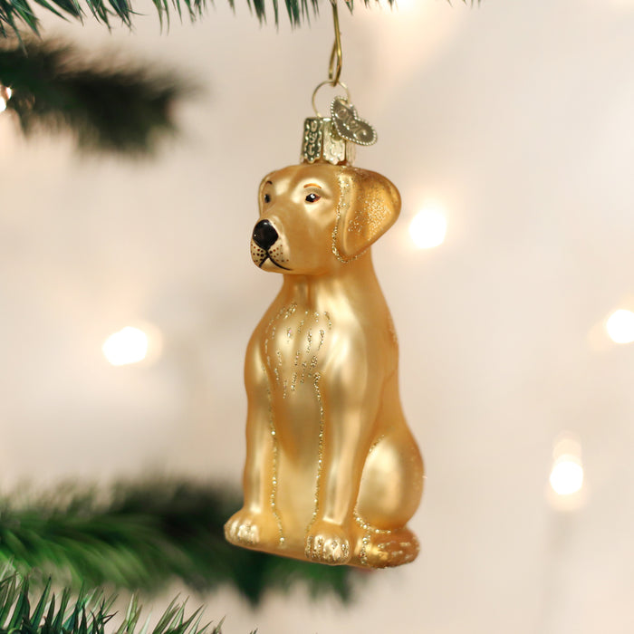 A golden dog-shaped Christmas ornament hangs from a tree branch, surrounded by blurred lights and green pine needles.