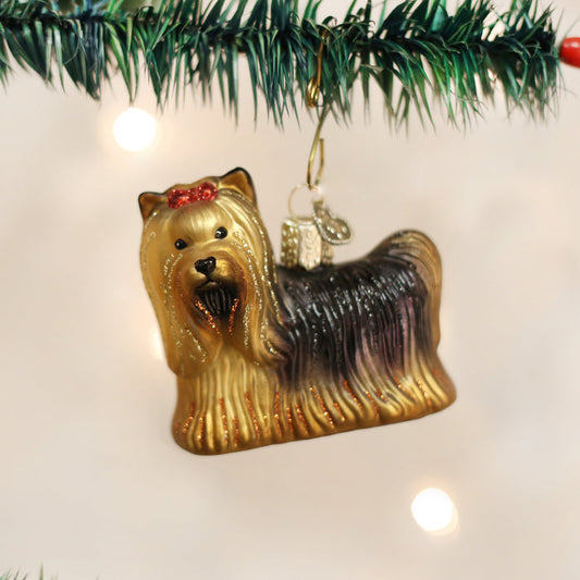 A shiny Christmas tree ornament shaped like a Yorkshire Terrier dog with a red bow is hanging from a green branch, with blurred holiday lights in the background.